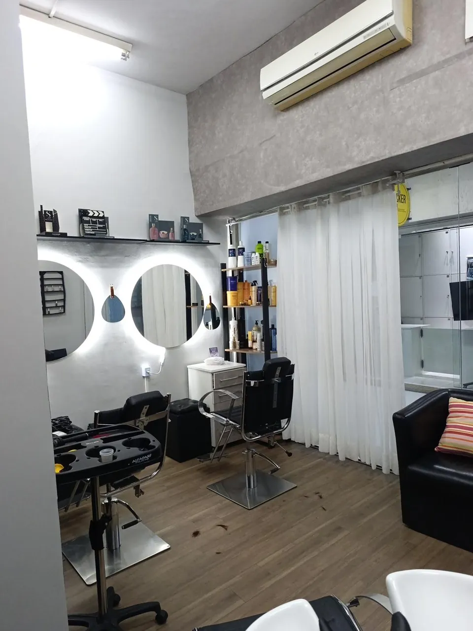 Interior do estúdio em Ipanema: cadeiras diante de espelhos redondos com luz, prateleiras e cortinas claras