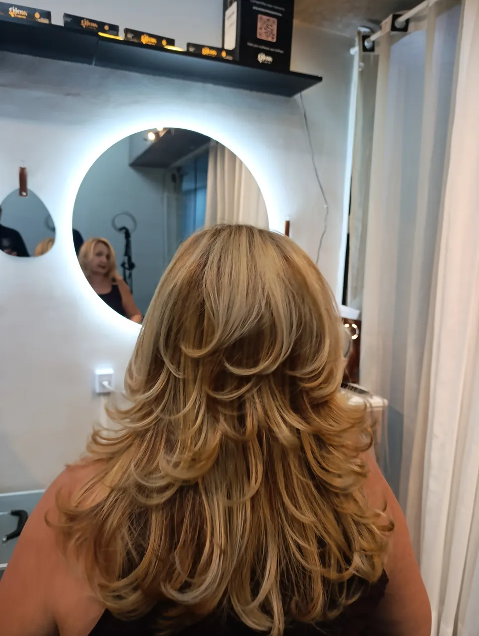 Espelho redondo iluminado do estúdio, com uma cliente de corte em camadas loiro de costas