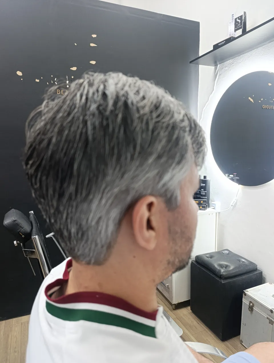 Corte masculino curto com grisalho natural, visto de lado, com espelho iluminado ao fundo