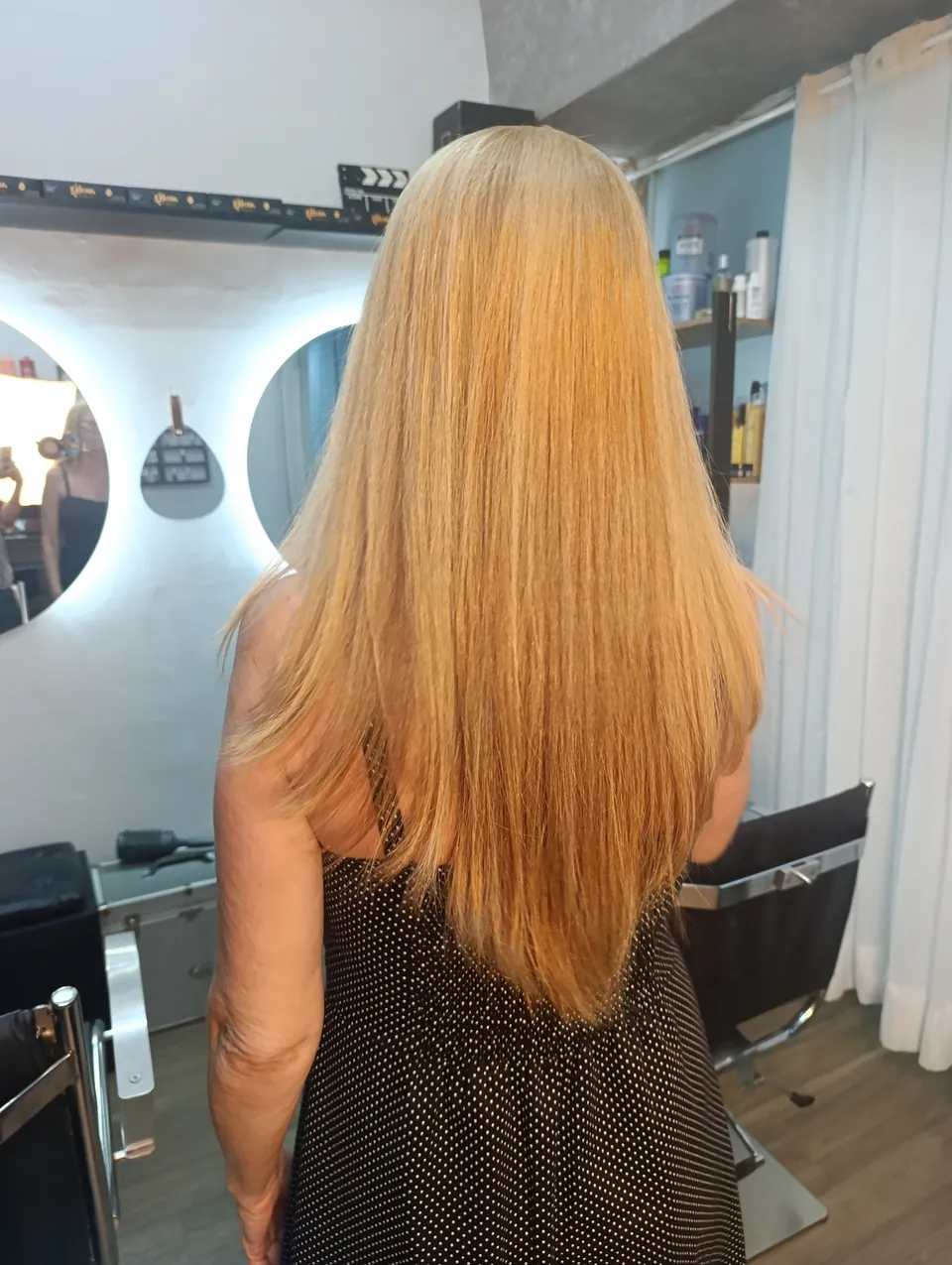 Cabelo longo e liso em loiro dourado, de costas, com espelhos iluminados ao fundo