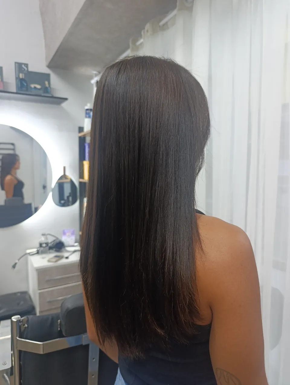 Cabelo castanho escuro longo e liso com brilho, de três quartos de costas, com espelho iluminado ao fundo