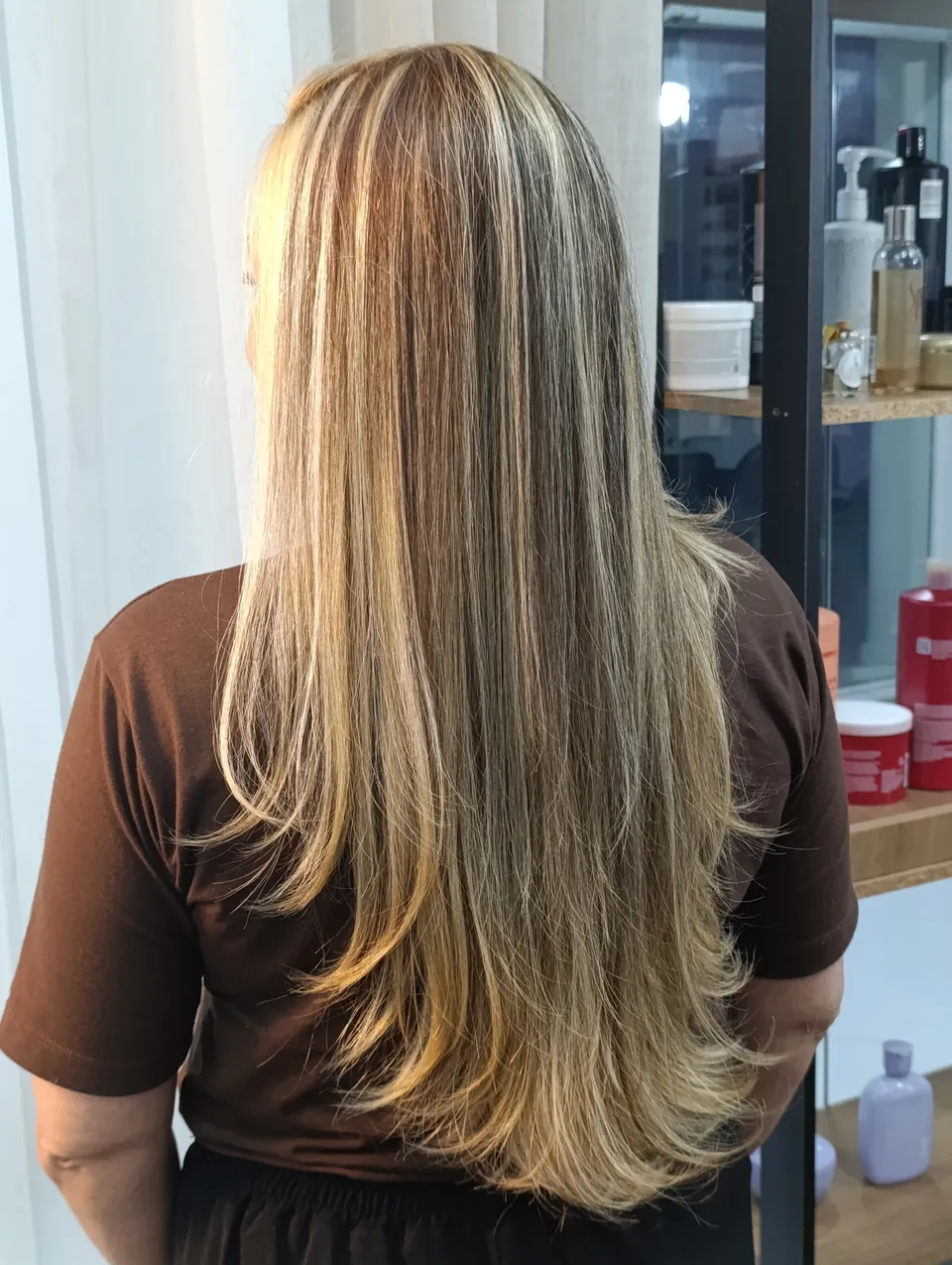 Liso longo com luzes loiras finas, de costas, com prateleira de produtos ao lado